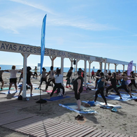 A group of people doing yoga on mats on a sandy beach in the sun, under a wooden shelter signed “Playas Accesibles de Vera”, with the sea and blue sky behind.