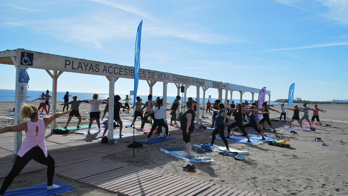 A group of people doing yoga on mats on a sandy beach in the sun, under a wooden shelter signed “Playas Accesibles de Vera”, with the sea and blue sky behind.