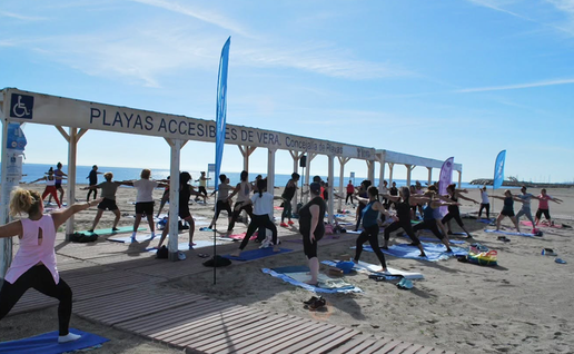 A group of people doing yoga on mats on a sandy beach in the sun, under a wooden shelter signed “Playas Accesibles de Vera”, with the sea and blue sky behind.