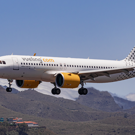 A white Vueling passenger jet with yellow engines and a dotted tail flying low over brown hills, with its landing gear down and a small village below.