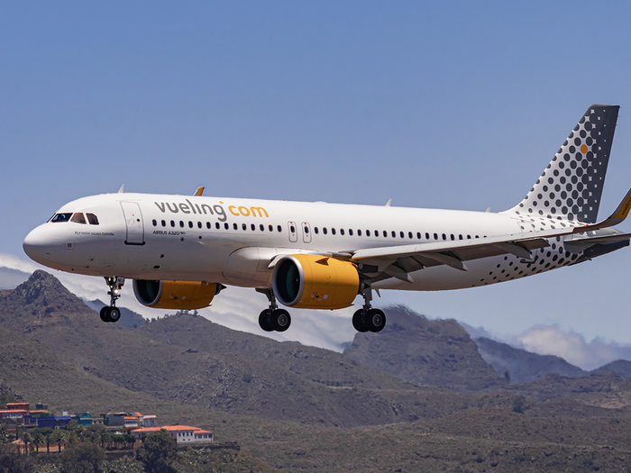 A white Vueling passenger jet with yellow engines and a dotted tail flying low over brown hills, with its landing gear down and a small village below.