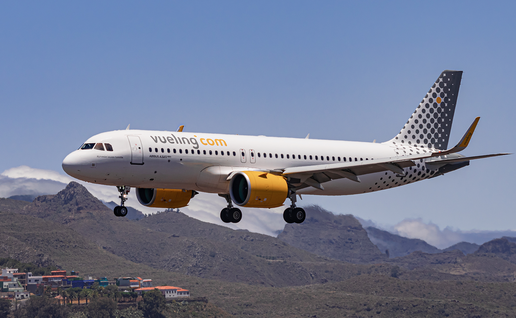 A white Vueling passenger jet with yellow engines and a dotted tail flying low over brown hills, with its landing gear down and a small village below.