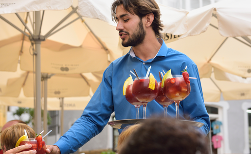 A waiter in a blue shirt serves glasses of sangria to customers at a café terrace.