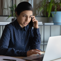 A woman in a navy polka-dot shirt holds a mobile phone to her ear while typing on a laptop at a desk with a notepad, paper and pen.
