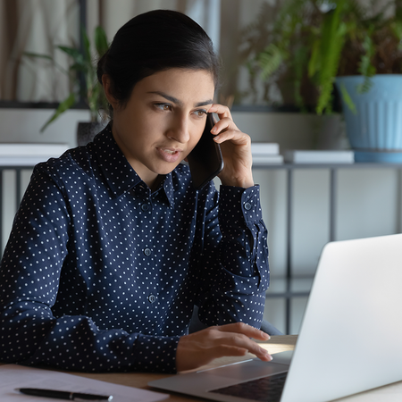 A woman in a navy polka-dot shirt holds a mobile phone to her ear while typing on a laptop at a desk with a notepad, paper and pen.