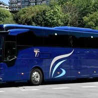 A dark blue BusBAM coach parked on a paved bay, with apartment blocks and trees behind it under a clear blue sky.