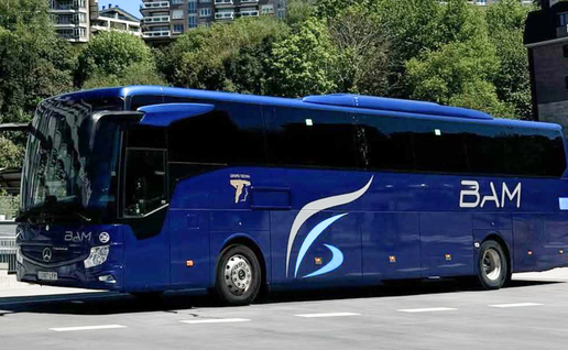 A dark blue BusBAM coach parked on a paved bay, with apartment blocks and trees behind it under a clear blue sky.