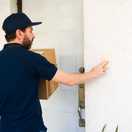 A man in a navy cap and polo shirt, carrying a cardboard box, presses the doorbell beside a white front door.