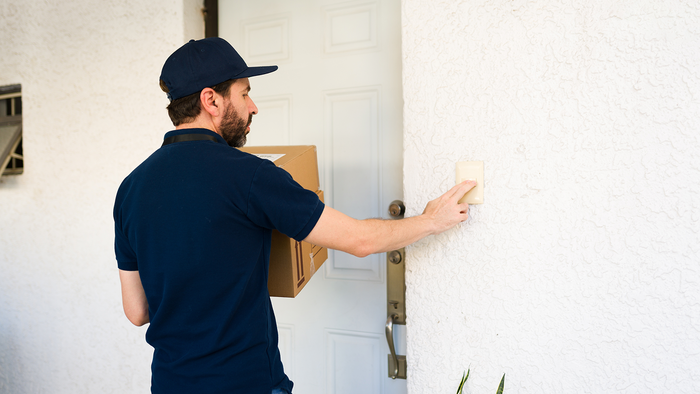 A man in a navy cap and polo shirt, carrying a cardboard box, presses the doorbell beside a white front door.