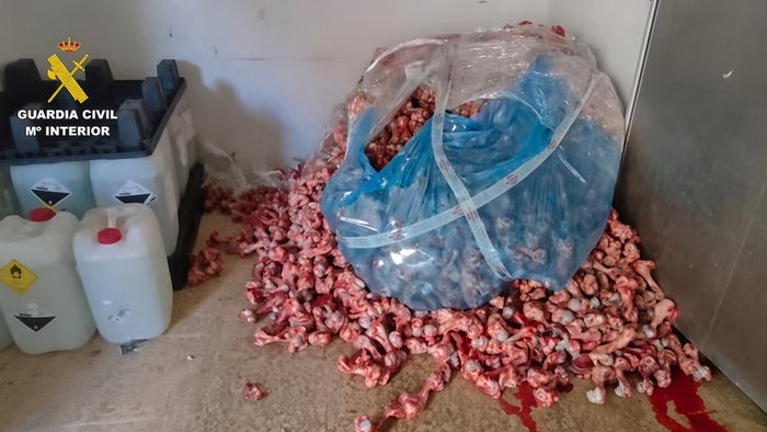 Pile of raw meat scraps and bones on a floor next to chemical containers and a blue plastic bag, with Guardia Civil logo in the top left.