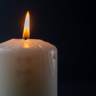 Alt Text: A single white pillar candle burning with a steady flame against a dark background, with wax drips visible along its side.