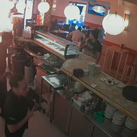 CCTV view of a restaurant kitchen and counter with staff, customers, and a bright fire or light flare on the left.
