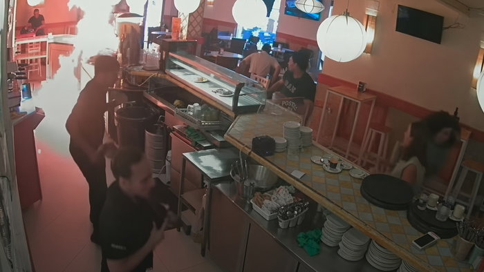 CCTV view of a restaurant kitchen and counter with staff, customers, and a bright fire or light flare on the left.