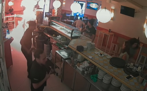 CCTV view of a restaurant kitchen and counter with staff, customers, and a bright fire or light flare on the left.